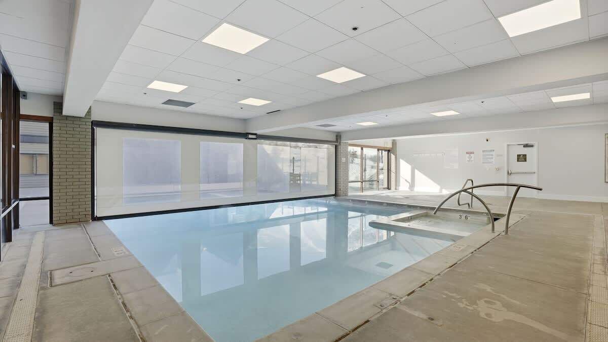 A large indoor swimming pool with clear glass walls, a tiled floor, and a ceiling with recessed lighting.