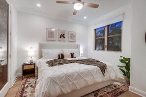 Une chambre avec un grand lit, un ventilateur de plafond et une fenêtre donnant sur des palmiers.