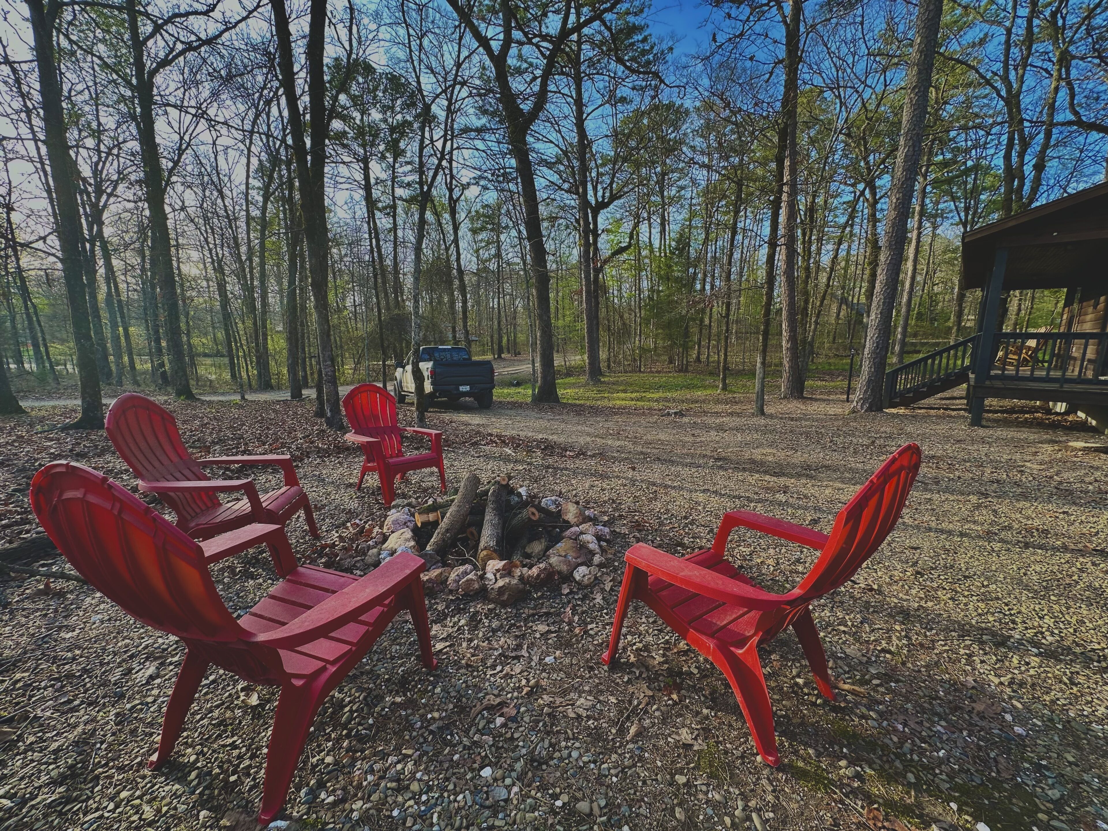 A campsite with red chairs arranged around a fire pit, surrounded by trees and a wooden deck.