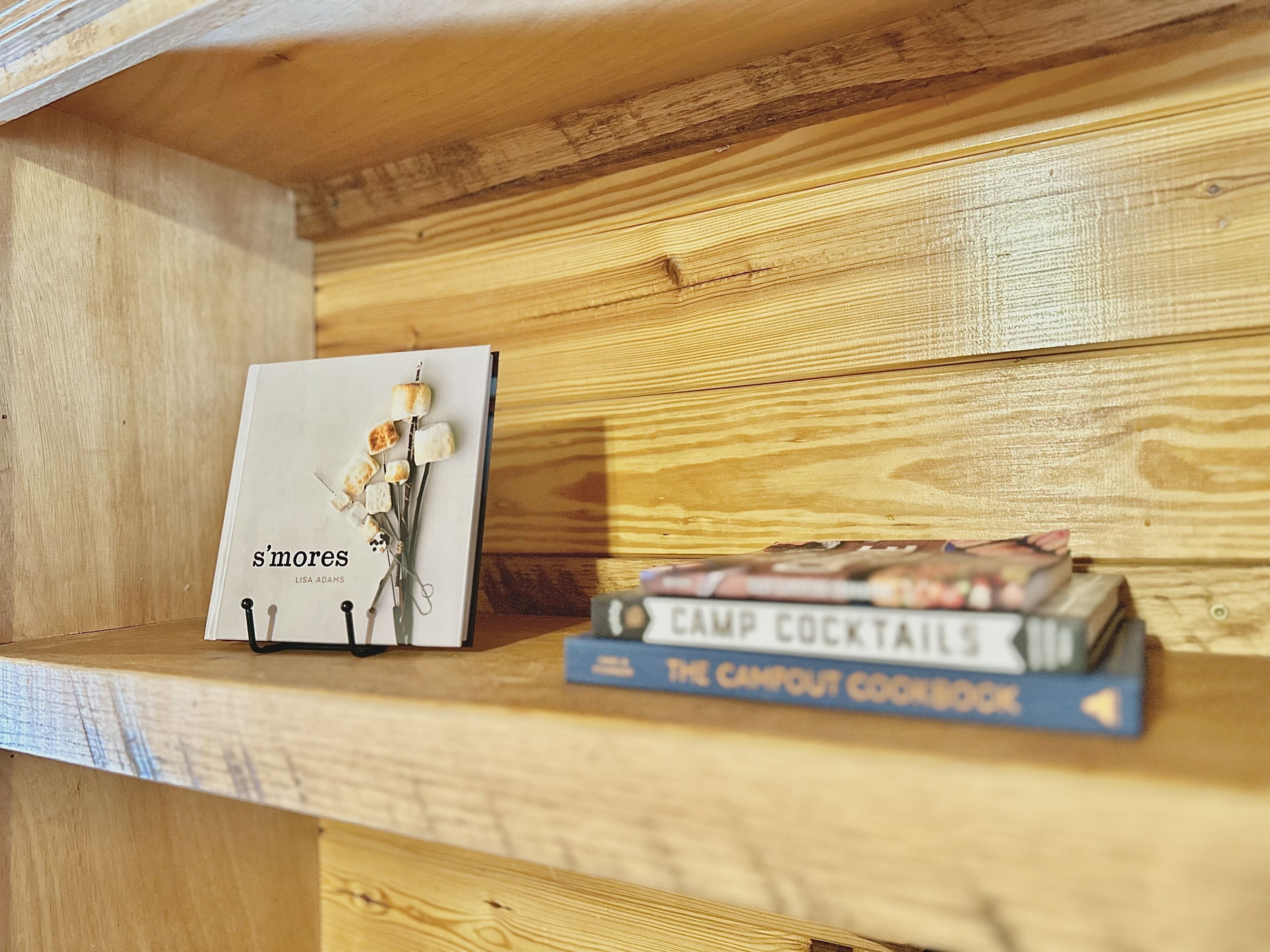A wooden shelf with a book titled 's'mores' by Lisa Adams and 'Camp Cocktails: The Campout Cookbook' by the same author.