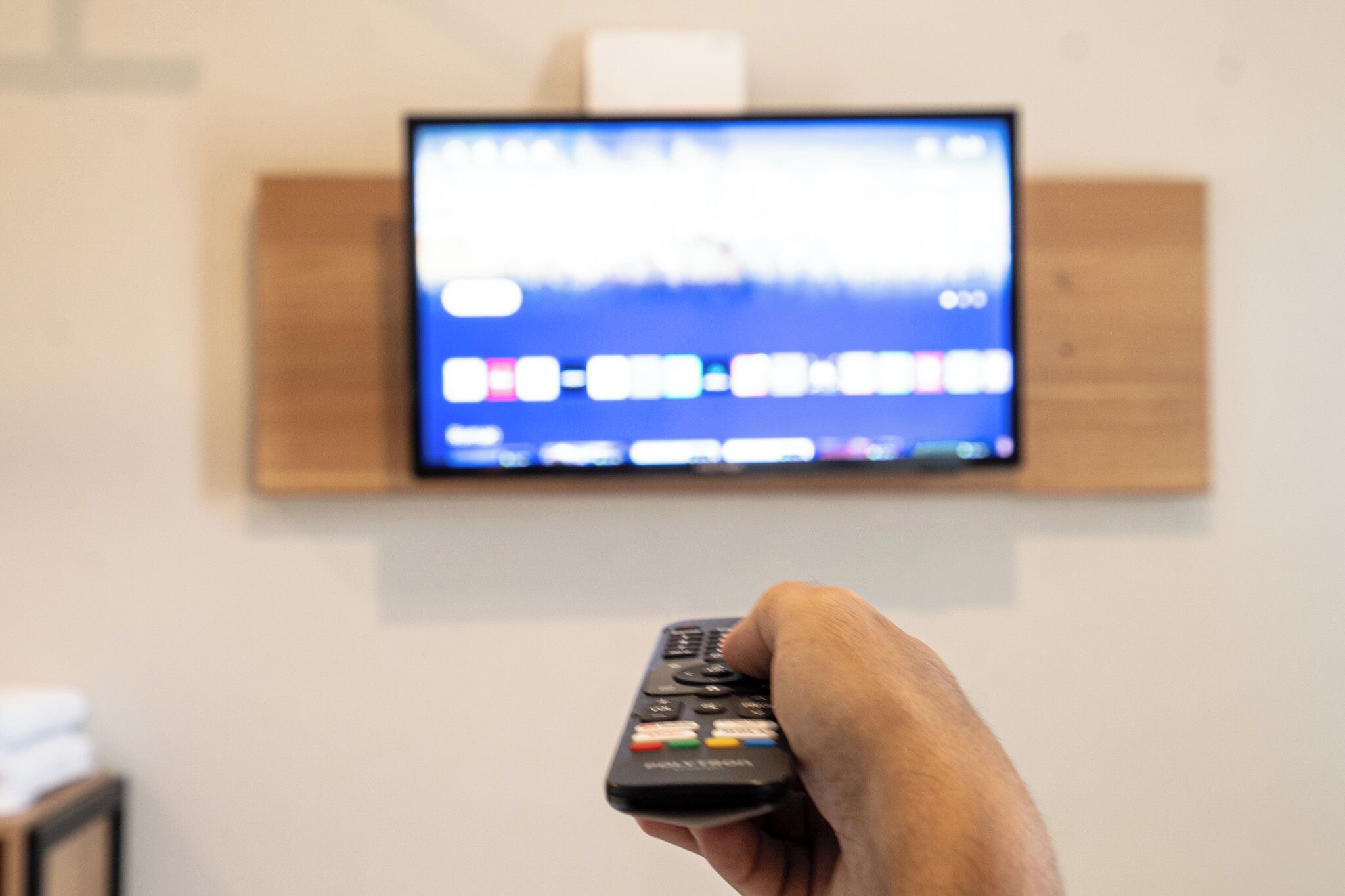 A hand holding a remote control in front of a television mounted on a wall.