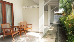 A covered outdoor seating area with wooden chairs and a table, adjacent to a building with a white exterior and a window with a wooden frame. - Maluk Stay (Sumbawa Barat)