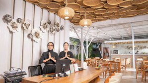 A modern interior with a wooden reception desk, two staff members, and a dining area with wooden tables and chairs. - Maluk Stay (Sumbawa Barat)
