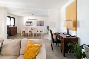 Een moderne woonkamer met een bank, een salontafel, een eethoek met tafel en stoelen, een houten bureau met lamp, en een groot schilderij aan de muur.