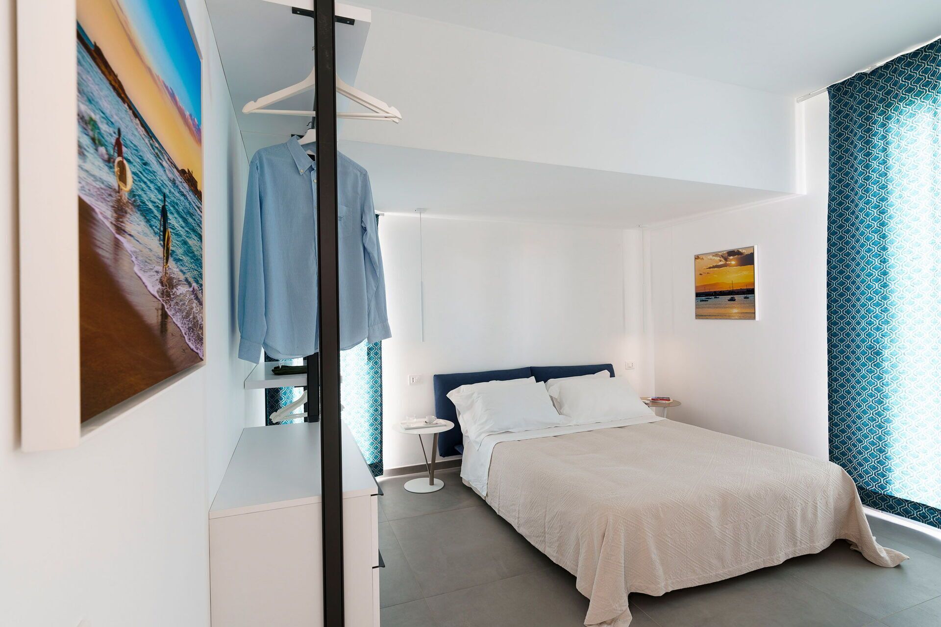 Una camera da letto con un letto, un comodino, una lampada a sospensione, una finestra con tende e un quadro appeso al muro.