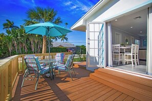 Een houten terras met buitenmeubilair, een turquoise parasol, en uitzicht op een huis met een witte gevel en een glazen deur die leidt naar een eethoek.