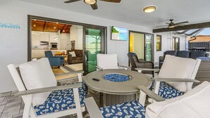 A patio area with a round table, white and blue patterned chairs, a fire pit, ceiling fans, and a view into the kitchen. - Anchors Away - Pool with Spa - Garage Game Room (Cape Coral)