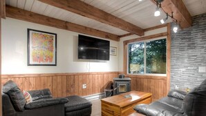 Un salon chaleureux avec des boiseries, une cheminée, un téléviseur et une grande fenêtre donnant sur les arbres.