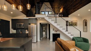 A modern kitchen with a central island, a staircase leading to an upper level, and a living area with a sofa and green armchair. - Secluded Montana A-Frame with Hot Tub and Views (Jefferson City)