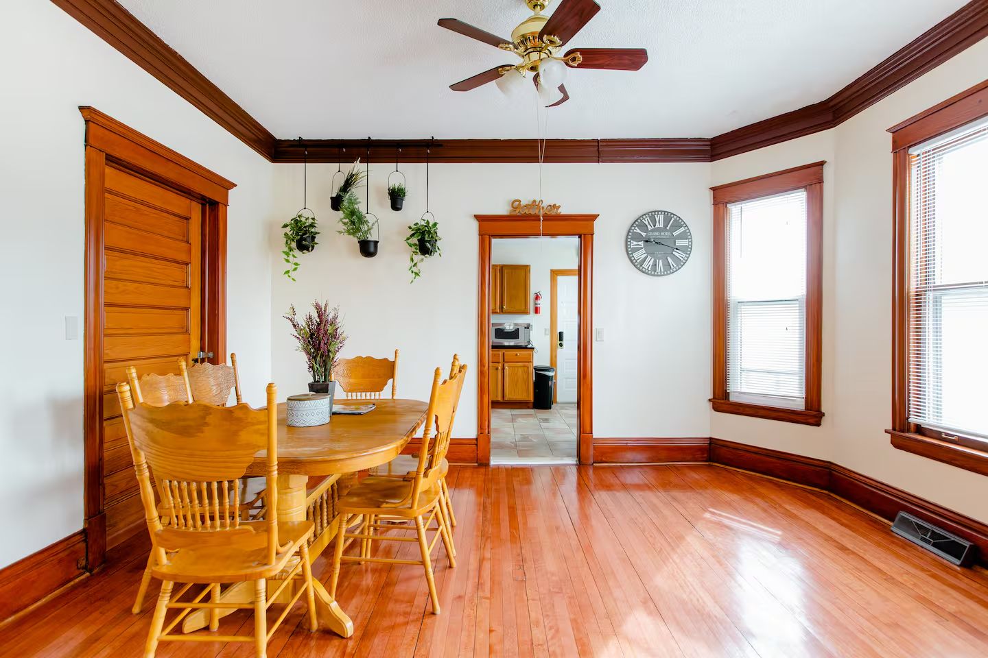 A dining room with a wooden table and chairs, a ceiling fan, hanging plants, and a clock on the wall.