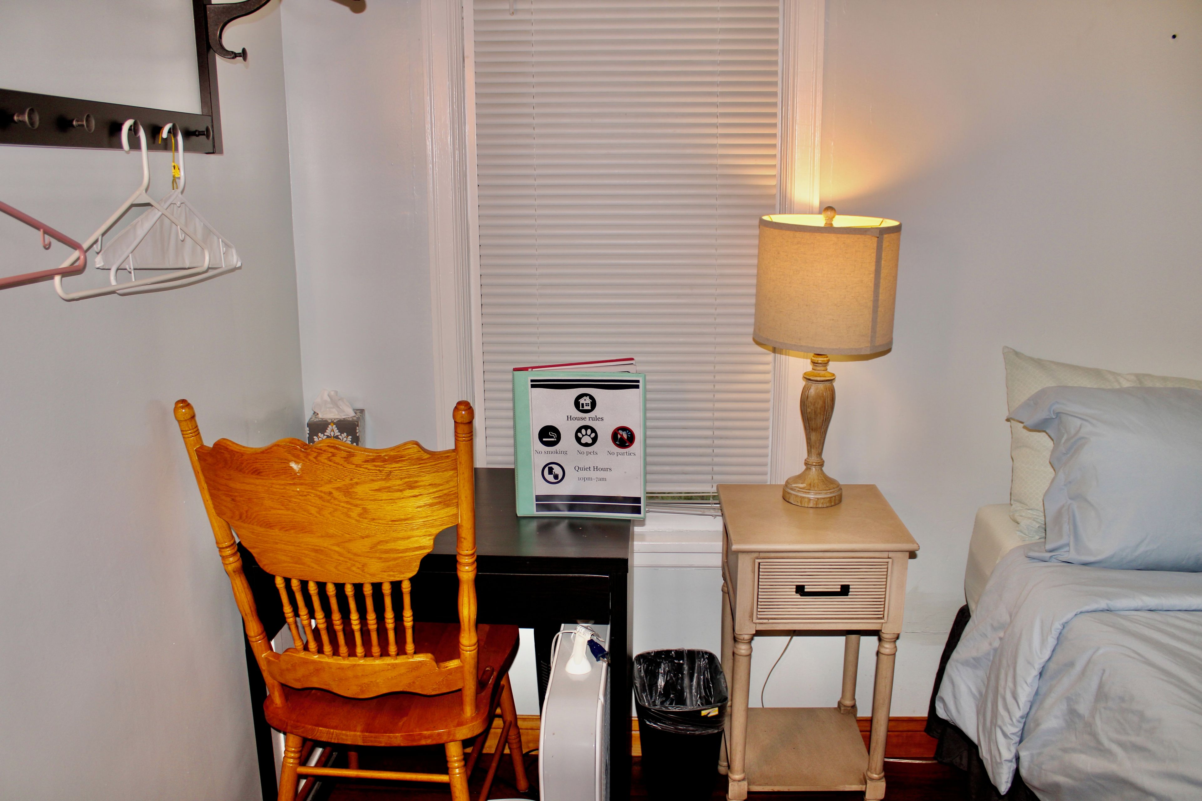Une chambre d’hôtel avec une chaise en bois, une table de chevet avec une lampe, un lit, une poubelle et un panneau indiquant le règlement intérieur.