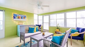 Una habitación con pared verde, ventilador de techo, ventana con vistas, mesa con sillas y sofá con cojines azules y amarillos.