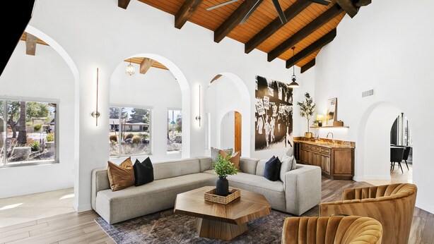 Uma sala de estar ampla com um sofá modular grande, uma mesa de centro de madeira e uma poltrona confortável. O espaço possui vigas de madeira aparentes, janelas grandes e vista para o exterior.