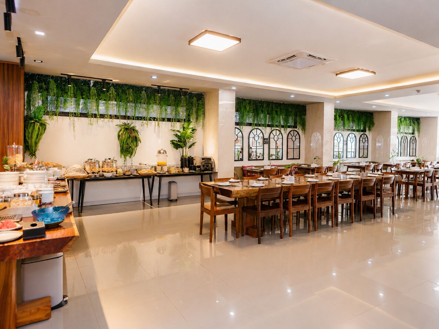 A spacious dining area with multiple tables and chairs, a buffet setup on the left, and large windows with decorative greenery.