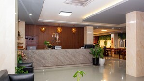 Zona de recepción de hotel con mostrador de mármol, escritorio de recepción, área de descanso y plantas en macetas.