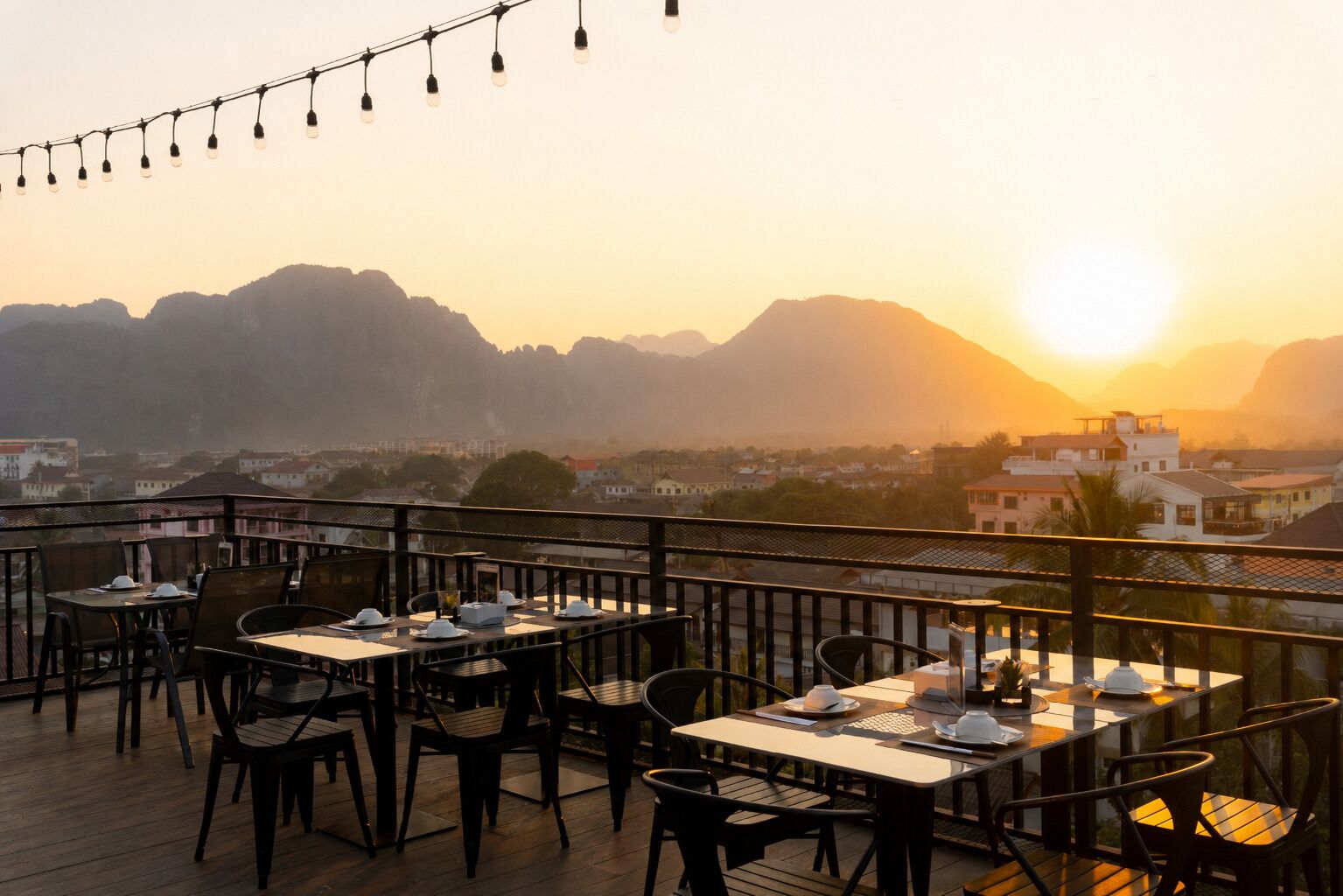 An outdoor dining area with tables and chairs, overlooking a cityscape and mountains during sunset.