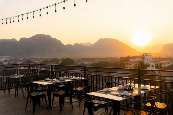 An outdoor dining area with tables and chairs, overlooking a cityscape and mountains during sunset.