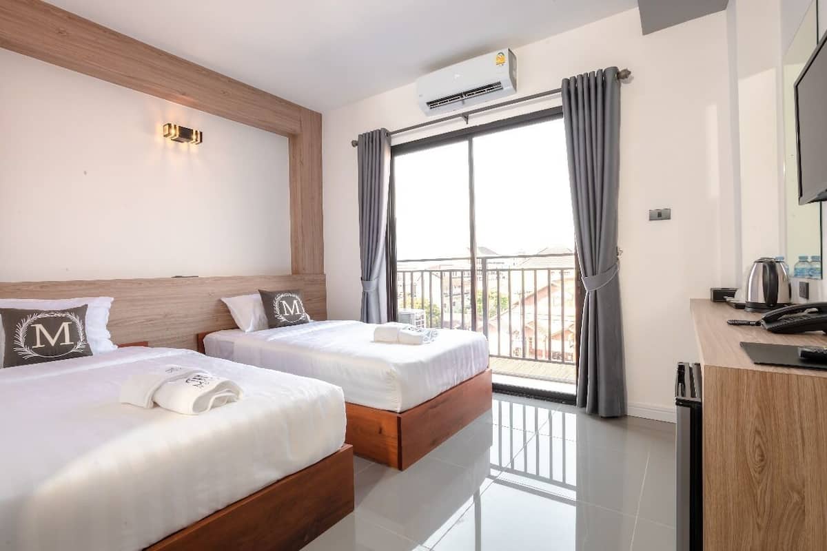 A hotel room with two beds, a wooden headboard, an air conditioning unit, a balcony with curtains, a desk with a phone, and a TV.