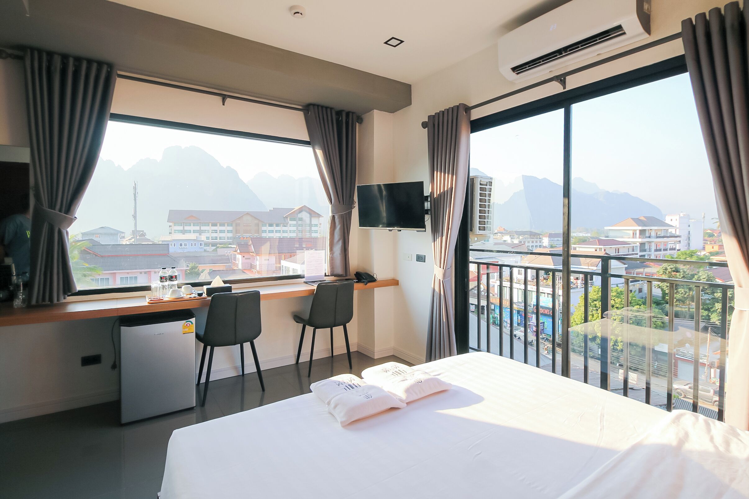 A hotel room with a bed, desk, chairs, a television, and a view of a cityscape.