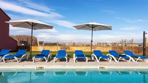 Een zwembadgedeelte met blauwe ligbedden en parasols, een helderblauwe lucht, en in de achtergrond een hek en bomen.