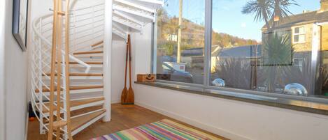 A room with a spiral staircase, a large window with a view of a house and palm tree, and a colorful striped rug on the floor.