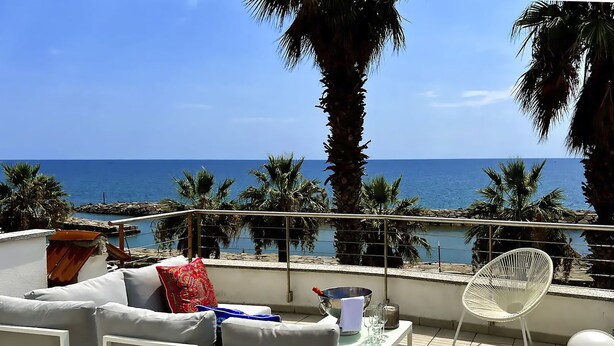 Una terraza con un sofá, una silla blanca y una mesa con una botella y vasos. La terraza tiene vistas a una playa con palmeras y el mar.