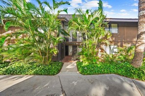 A modern two-story building with a concrete pathway leading to the entrance, surrounded by lush greenery and palm trees. - Remodeled Beachfront 2BD w Year Round Beach - Maalaea Surf A6 (Kihei)