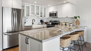 A modern kitchen with white cabinetry, stainless steel appliances, a central island with bar stools, and a decorative plant. - Remodeled Beachfront 2BD w Year Round Beach - Maalaea Surf A6 (Kihei)