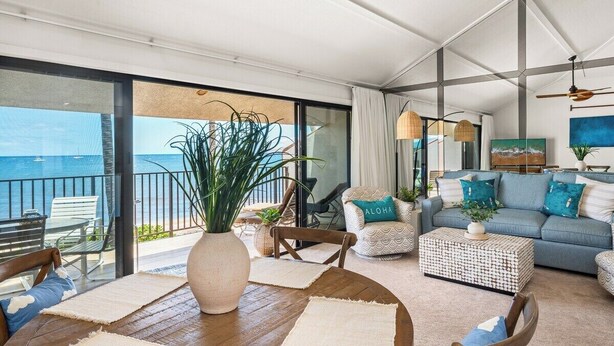 A spacious living area with a large window overlooking the sea, a wooden dining table, and a comfortable seating arrangement. - Remodeled Beachfront 2BD w Year Round Beach - Maalaea Surf A6 (Kihei)