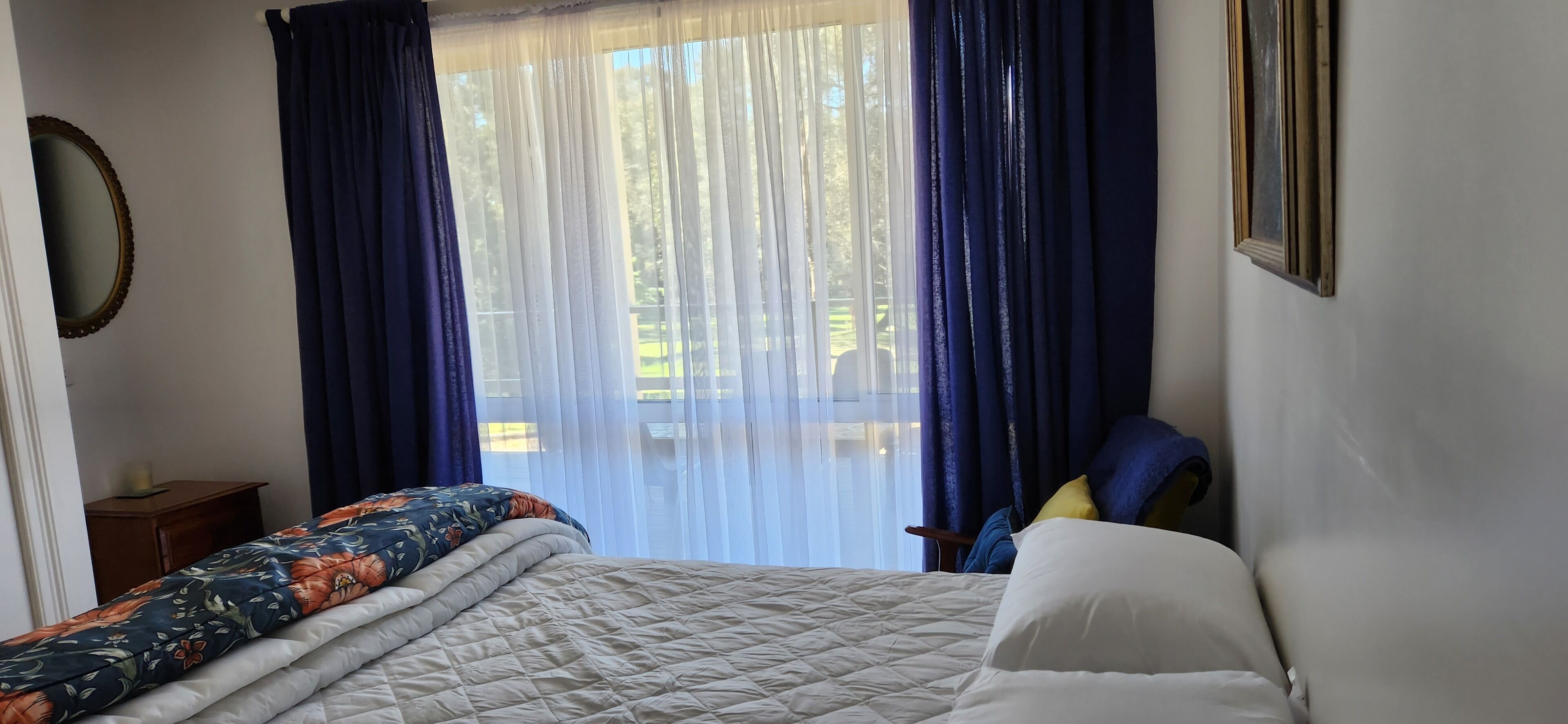 Ein Schlafzimmer mit Bett, Nachttisch, Spiegel und Blick ins Freie durch transparente Vorhänge.