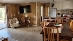 Ein gemütlicher, gut beleuchteter Raum mit Holzboden, einem großen an der Wand montierten Fernseher, einem Esstisch mit Stühlen und einer kleinen Küchenzeile mit Kühlschrank und Herd.
