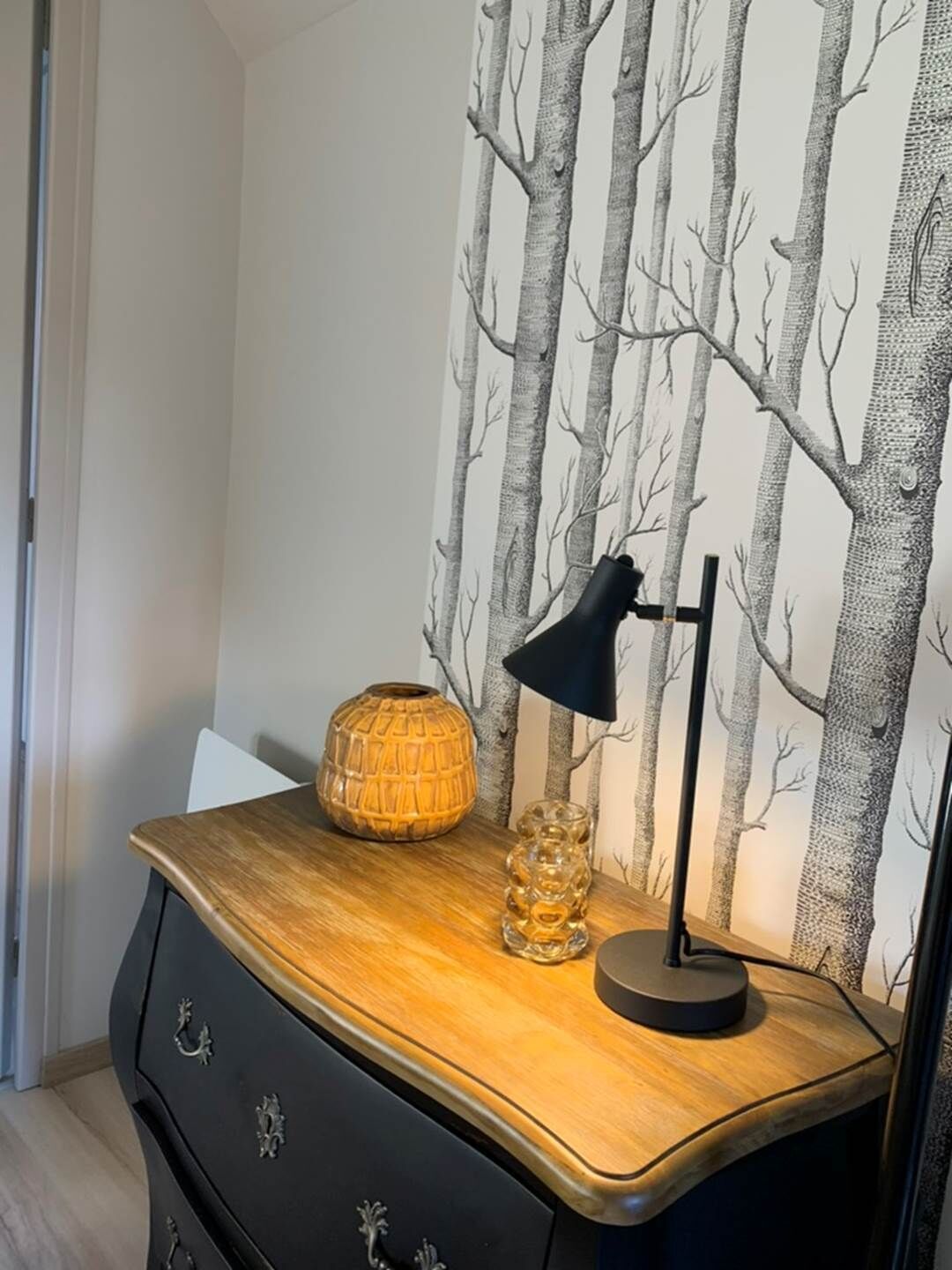 Un escritorio de madera con una lámpara, un florero texturizado y una pared con estampado de árbol.