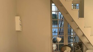 Una sala de estar acogedora con un sofá, una mesita redonda y una silla. En el fondo, se ve una escalera con un ventanal.