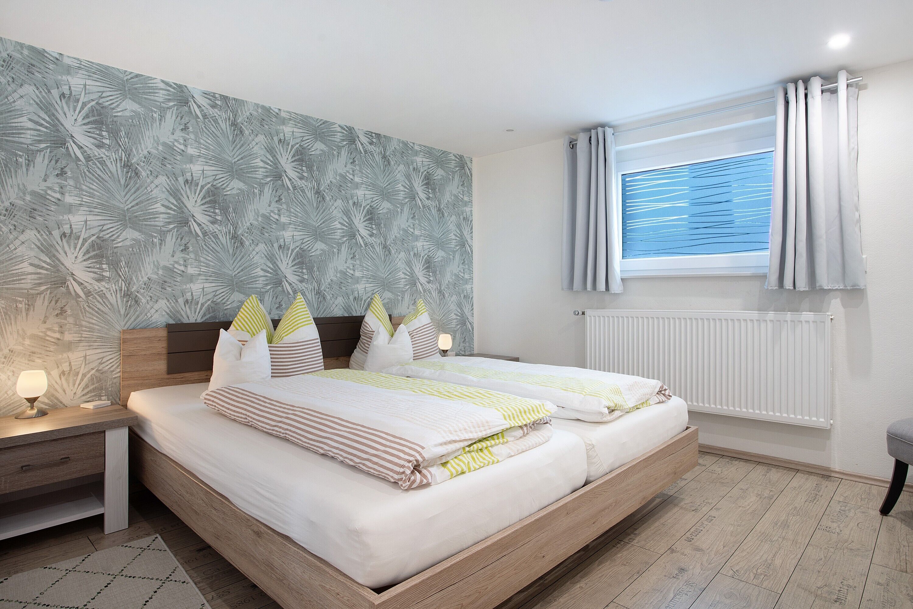 Ein Doppelbett mit Holzrahmen, ein Nachttisch mit Lampe, ein Fenster mit Vorhängen und eine Heizung.