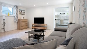 Un salon moderne avec un canapé, une table basse, un téléviseur posé sur un meuble en bois, et une vue sur la cuisine.