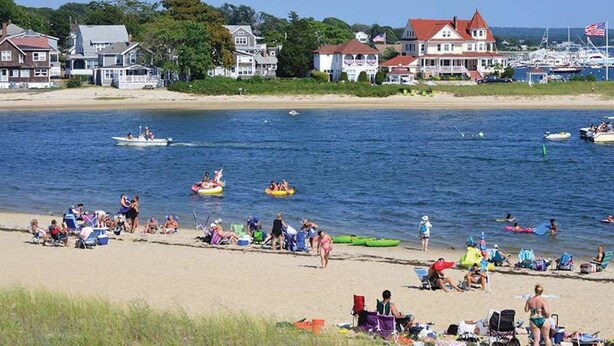 A beach with people sunbathing and swimming, boats on the water, and houses in the background. - Modern Beachside Condo in Onset Village: Walk to Dining, Market & Beaches Monthly Rates Available (Onset)