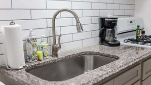A kitchen with a stainless steel sink, white tiled backsplash, a coffee maker, and a gas stove.