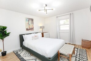 Un dormitorio con una cama, una mesita de noche, una lámpara, una ventana con cortinas, una planta y una cesta tejida.