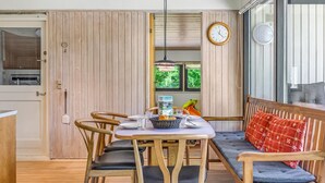 Un comedor con mesa y sillas de madera, un banco con cojines, un reloj en la pared y una ventana con follaje en el exterior.