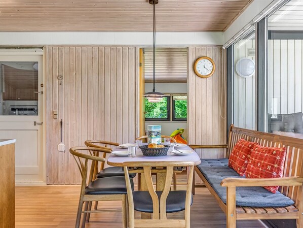 A dining area with a wooden table and chairs, a bench with cushions, a clock on the wall, and a window with greenery outside.