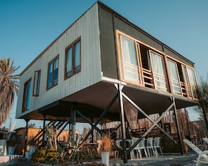 Ein zweistöckiges Gebäude mit einem Balkon, das von Metallträgern gestützt wird und von verschiedenen Pflanzen und Gartenmöbeln umgeben ist.