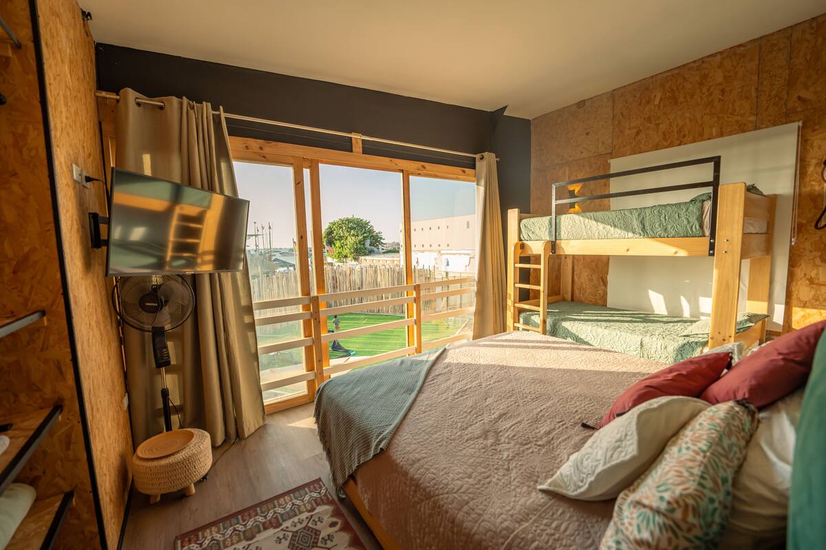 Quarto com beliche, ventilador, janela com cortinas e vista para uma paisagem verde.
