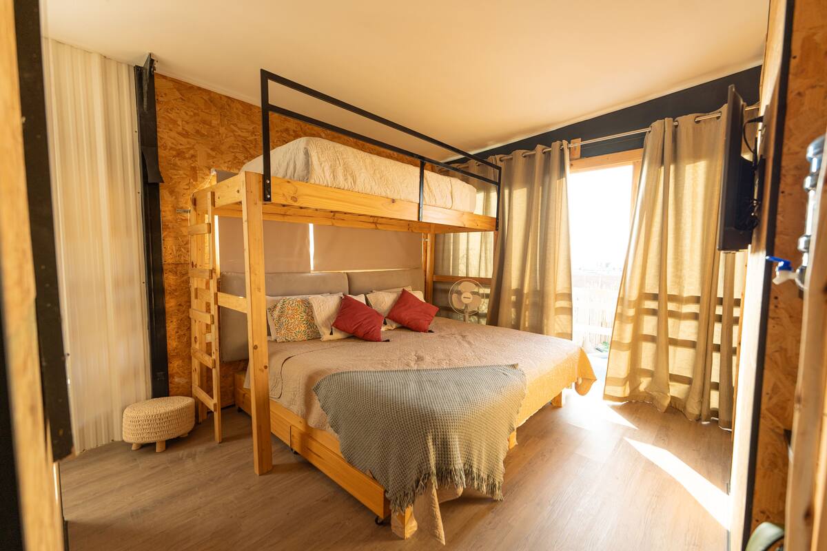 Quarto com beliche, cabeceira de madeira, um pequeno banquinho e janela com cortinas.