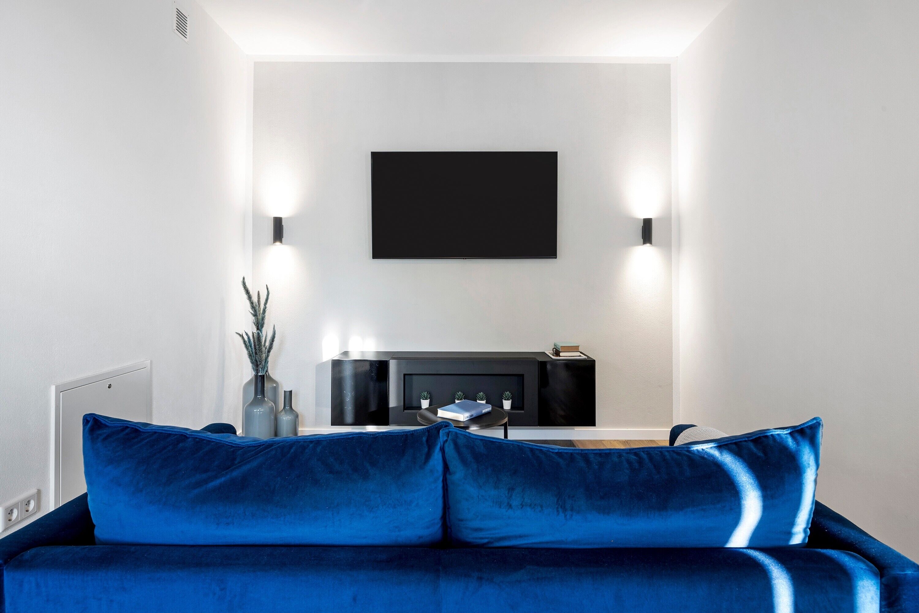 Un salon moderne avec un canapé bleu, un meuble de rangement noir pour les appareils électroniques et des lampes fixées au mur.