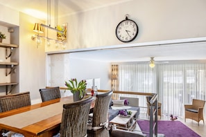 Ein modernes Wohnzimmer mit Esstisch, Korbstühlen, einer Couch, einer Uhr und einem großen Fenster mit Vorhängen.