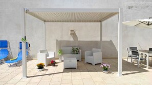 A covered outdoor seating area with white wicker furniture, a blue lounge chair, and a table with chairs.
