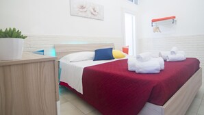 A hotel room with a bed, a bedside table, a potted plant, a red bedspread, and a white wall with a picture. - Casa Camerini in Villanova (Ostuni)