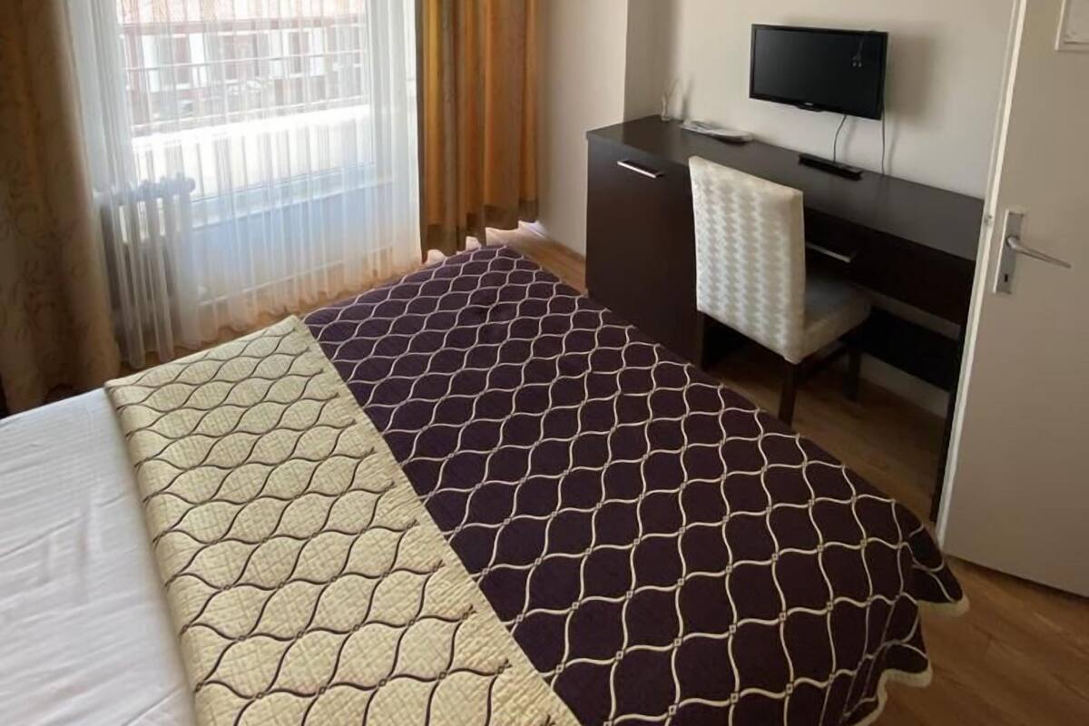 Quarto de hotel com cama, escrivaninha, televisão e ventoinha no teto.