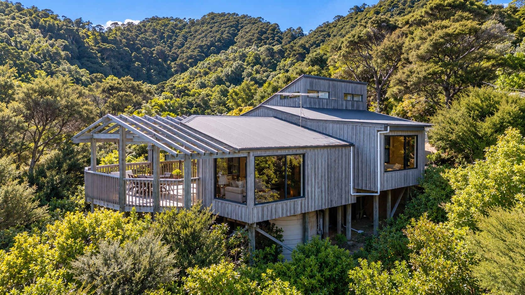 A modern wooden house with a deck, large windows, and a view of a forested hillside.