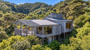 A modern wooden house with a deck, large windows, and a view of a forested hillside.